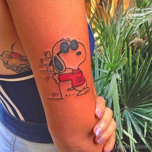 Фото татуировки с изображением Снупи из мультфильма "Peanuts" в красной футболке, расположенной на мужском предплечье.