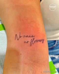 Фотография минималистичной татуировки с надписью "No rain no flowers" на руке. Татуировка выполнена тонкими линиями.