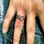 Фотография миниатюрной татуировки с надписью "Vici" на пальце руки. Черный цвет, аккуратный шрифт.