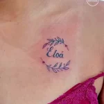 Фото тату: небольшая татуировка с именем "Eloá" и ветками с цветами, выполненная на ключице.