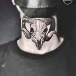Фото тату черепа барана на шее. Детализированный рисунок с проработанными рогами и глазницами. Черно-белая татуировка в стиле реализм.