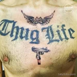 Фотография татуировки с надписью 'Thug Life', пистолетом и короной на груди мужчины. Рисунок выполнен в черно-белых тонах.