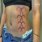 Фотография минималистичной черно-белой татуировки на боку девушки. Рисунок состоит из нескольких тонких линий, образующих абстрактный узор.