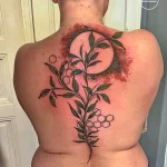 Фотография татуировки полумесяца с ветками, листьями и сотами на спине девушки. Рисунок выполнен в сочетании черного и красного цветов.