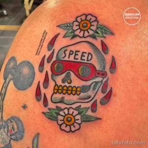 Фотография олдскульной татуировки черепа в очках и с надписью 'SPEED' на плече. Рисунок выполнен в традиционном стиле с использованием ярких цветов и четких контуров.