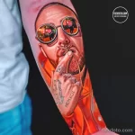 Фотография яркой татуировки с изображением рэпера Мак Миллера на руке. Детализированный портрет, очки, цепи и надпись 'MCMXCII'.