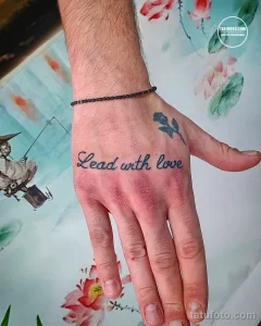 Фото тату: надпись "Lead with love" и маленький цветок на руке, выполненные в акварельной технике.