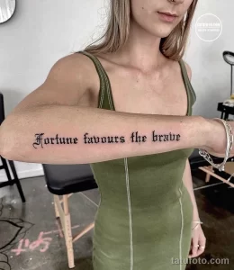 Фото тату надписи 'Fortune favours the brave' на предплечье. Черный шрифт в классическом стиле, четкие линии, минималистичный дизайн.