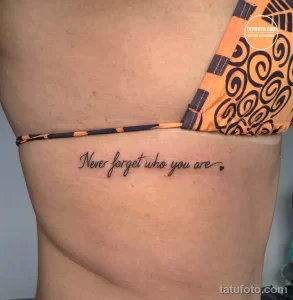 Фотография татуировки на боку девушки с надписью 'Never forget who you are' и изображением сердца. Татуировка выполнена тонким шрифтом.