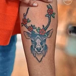 Фото тату: миниатюрный рисунок головы оленя с рогами, украшенными цветами, на руке. Яркая, цветная татуировка в акварельном стиле.