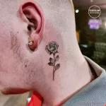 Фото тату: маленькая черная роза с листьями на шее девушки. Тонкие линии, реалистичное изображение, минималистичный стиль. Изящный дизайн.