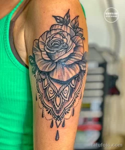 Фото татуировки с изображением розы, заключенной в сложный узор мандалы на плече. Использованы оттенки серого и черного для создания детализированного рисунка.