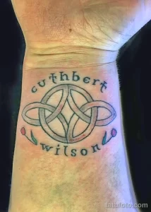 Фото тату: татуировка кельтского узла с надписью "cuthbert wilson" на запястье. Рисунок выполнен в черном цвете с добавлением красных акцентов.