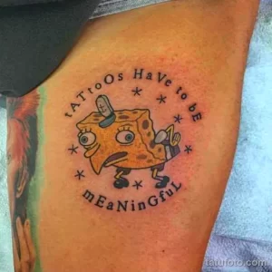 Фото тату Губки Боба на плече с надписью 'Tattoos Have to Be Meaningful'. Яркий, мультяшный стиль, юмористический рисунок татуировки.