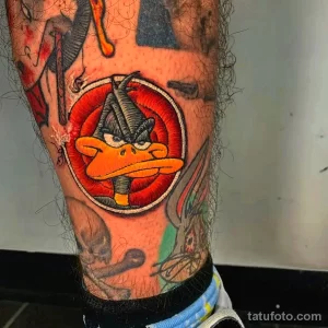 Фото тату: яркая татуировка Дакки Дака из Looney Tunes в красном круге на ноге, выполненная в мультяшном стиле.