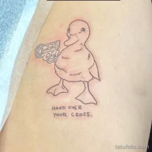 Фото тату: утка с пистолетом и надписью "HAND OVER YOUR CROCS", выполненная в черном цвете. Расположение - предплечье.