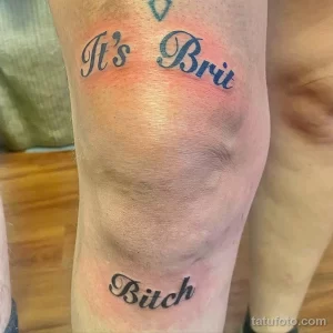 Фото тату: татуировка с надписью "It's Brit Bitch" в каллиграфическом стиле, выполненная синим цветом на ноге.