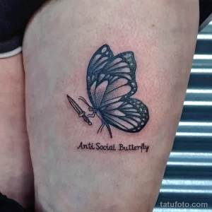 Фото тату: изображение бабочки, держащей в лапках кинжал, с надписью 'Anti Social Butterfly'. Черно-белая татуировка в стиле дотворк.