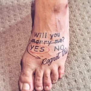Фото тату на стопе с надписью 'Will you marry me? YES NO Regret this'. Необычный и романтичный рисунок, выполненный в каллиграфическом стиле.