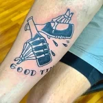 Фото тату на ноге с изображением скелета, держащего бутылку, и надписью "Good Times". Стильный и символичный рисунок.