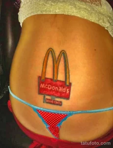 Фото тату на пояснице: логотип McDonald's с золотыми арками и надписью, выполненный в ярких цветах.
