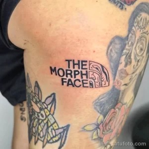 Фото минималистичной татуировки с надписью 'The Morph Face' и логотипом The North Face. Черный цвет, тонкие линии, плечо.