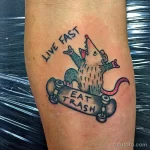 Фото тату крысы, катающейся на скейтборде, с надписью 'Live Fast Eat Trash'. Стиль олдскул, яркие цвета, предплечье.