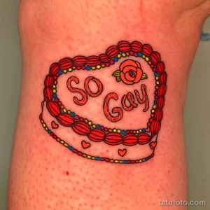 Фото тату: красочная татуировка с надписью "So Gay" и изображением сердца, украшенного цветами и узорами на руке.