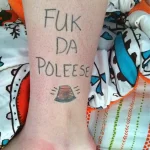 Фото тату: надпись "F*ck Da Police" и изображение полицейской фуражки, выполненное в черном цвете, протестный рисунок.