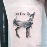 Фото тату олененка с надписью "Dill Doe" и блестками на руке. Рисунок татуировки выполнен в минималистичном стиле с использованием черного цвета.