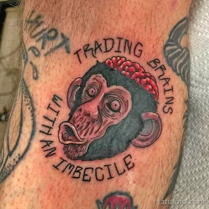 Фото тату обезьяны с вываливающимся мозгом и надписью "Trading Brains With An Imbecile" на плече. Рисунок татуировки выполнен в стиле традиционной татуировки.
