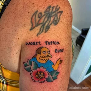 Фото тату на плече с изображением Гомера Симпсона и надписью 'WORST. TATTOO. EVER!', выполненное в ярких цветах.