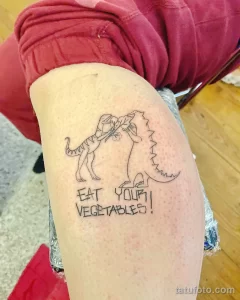 Фото тату на ноге с изображением динозавра, держащего овощи, и надписью 'EAT YOUR VEGETABLES!' в мультяшном стиле.