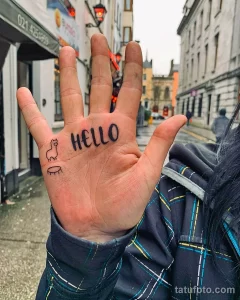 Фото тату на руке с изображением маленькой черной кошки и надписи 'HELLO', выполненной в простом шрифте. Фон - городской пейзаж.