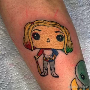 Яркая татуировка на руке с изображением Харли Квинн в стиле Funko Pop. Мультяшный стиль рисунка татуировки.