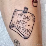 Фото тату, изображающего клочок бумаги с надписью "MY DAD HATES MY TATTOOS", прикрепленный к коже.