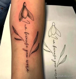 Черно-белое фото татуировки подснежника с надписью 'I am because you were' на предплечье. Изящный рисунок с тонкими линиями и каллиграфическим шрифтом.