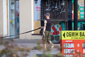 Фотография мужчины с татуировкой на руке, выгуливающего собаку на городской улице. Акцент на татуировке и повседневной обстановке.