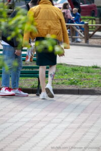 Фотография татуировки на ноге девушки в юбке, сидящей на скамейке в парке. Видны детали рисунка и окружающая обстановка.