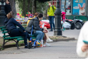 Фотография татуировки с изображением красной розы на ноге девушки, сидящей на скамейке в парке. Видны детали рисунка и окружающая обстановка.