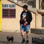 Фотография мужчины с татуировками на ногах, выгуливающего маленькую собаку на улице. Видны детали татуировок и общий городской пейзаж.