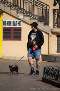Фотография мужчины с татуировками на ногах, выгуливающего маленькую собаку на улице. Видны детали татуировок и общий городской пейзаж.