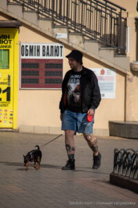 Фото тату на ногах мужчины, выгуливающего маленькую собачку на поводке в городской среде. Видны детализированные рисунки татуировок на голенях.