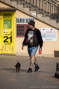 Фото мужчины с татуировками на ногах, выгуливающего маленькую собаку на фоне городской архитектуры. Идеи для тату и уличный стиль.