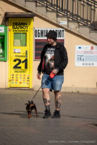Фото мужчины с татуировками на ногах, выгуливающего маленькую собаку на фоне городской архитектуры. Идеи для тату и уличный стиль.