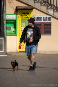 Фото мужчины с татуировками на ногах, выгуливающего маленькую собаку на фоне ярких городских зданий. Идеи для тату и уличный стиль.