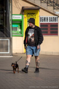 Фото мужчины с татуировками на ногах, выгуливающего маленькую собаку на улице города. Идеи для тату и уличный стиль.