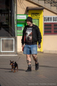 Фото мужчины с татуировками на ногах, выгуливающего маленькую собаку на фоне городской архитектуры. Идеи для тату и уличный стиль.