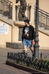 Фотография мужчины с татуировками на ногах, выгуливающего собаку возле здания. Видны детали татуировок и общий городской пейзаж.