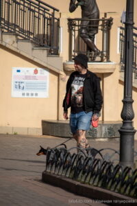 Фото тату на ногах мужчины, выгуливающего маленькую собаку на улице. Видны детализированные рисунки татуировок на голенях и бедрах.